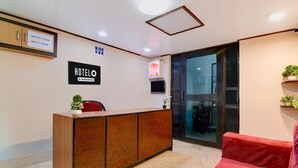 Un espace d’accueil d’hôtel avec un comptoir de réception en bois, un canapé rouge et un panneau indiquant les heures d’enregistrement.