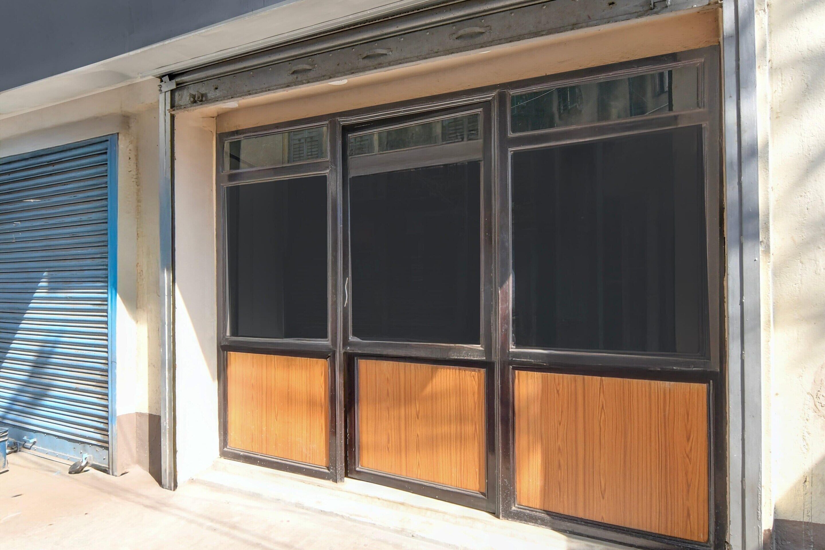 A building facade with a large window with a black frame and wooden panels, a blue shutter door, and a concrete ground.