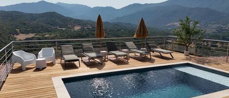 A poolside area with lounge chairs and umbrellas, overlooking a mountainous landscape.
