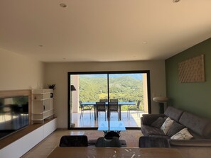 A modern living room with a sliding glass door leading to an outdoor patio, a flat-screen TV, a sofa, and a dining area with chairs.