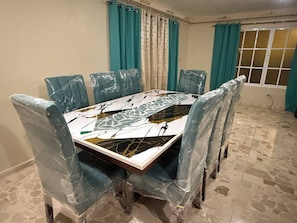A dining table with chairs wrapped in plastic, a tiled floor, and teal curtains. - Segundo Piso Completó (Pedro Brand)