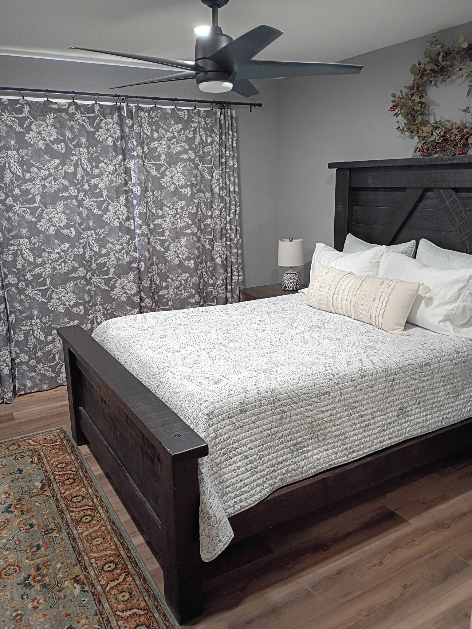 A bedroom with a dark wooden bed, a ceiling fan, a floral curtain, a bedside lamp, and a decorative wreath on the wall.