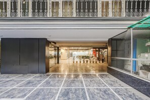Un hall espacioso con piso de mármol, barandilla metálica y puerta de vidrio que da a un restaurante.
