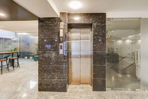 Un ascensor en un vestíbulo con pared de mármol, puerta de cristal y una zona de estar con taburetes.