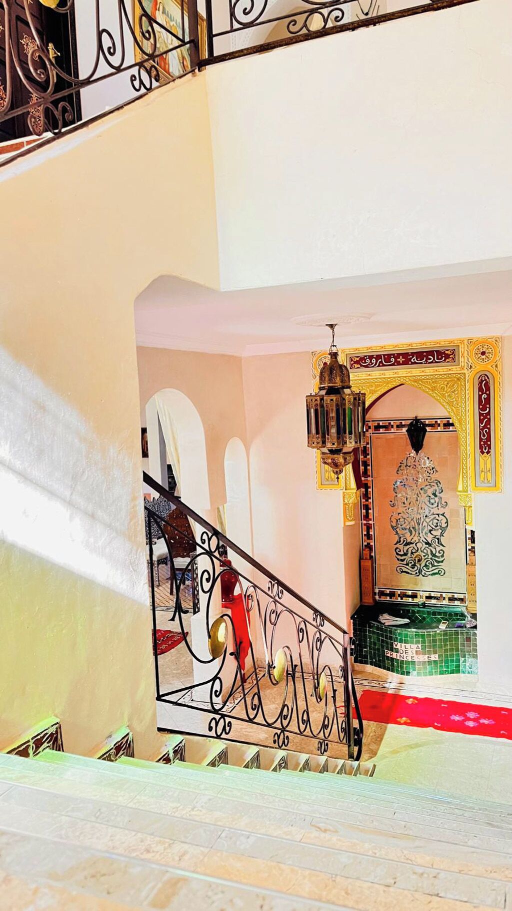 Un escalier orné de rampes en métal ouvragé, un lustre et une arche décorative agrémentée de calligraphie arabe.