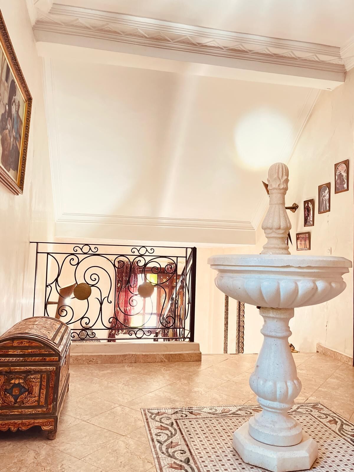 Une pièce avec une fontaine blanche, un tapis à motifs, un coffre décoratif et un escalier doté d’une rampe en fer forgé.
