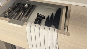 Una cocina con tope de inducción negro, utensilios de madera, un recipiente metálico para utensilios, dispensador de toallas de papel y un cajón con utensilios ordenados.