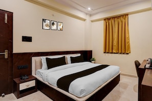 A hotel room with a bed, a desk, a chair, a plant, and artwork on the wall.