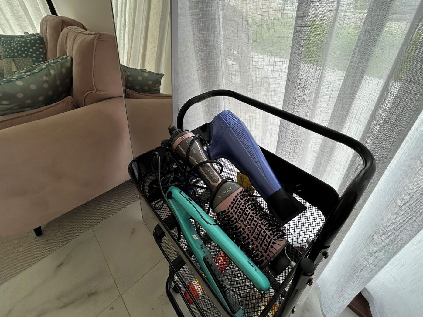 A room with a sofa, a window with sheer curtains, and a black mesh basket containing hair styling tools.