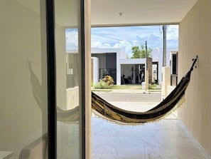 A modern interior with a hammock on a tiled floor, a glass door leading outside, and a clear view of the street and buildings. - House with Pool • 10min French • Cond. Closed (Marechal Deodoro)