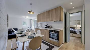 A modern kitchen with wooden cabinets, a dining area with a table and chairs, and a view into a bedroom with a bed.