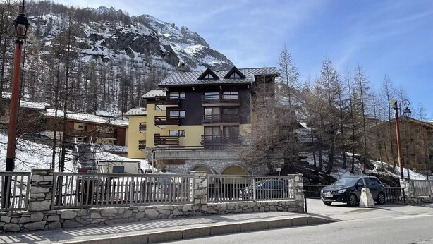 Un edificio de varios pisos con balcones, fachada de piedra y montañas al fondo.