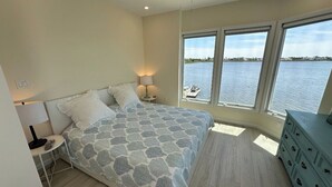 Ein Schlafzimmer mit Bett, Nachttischen, einem Schminktisch und Blick auf ein Gewässer durch große Fenster.
