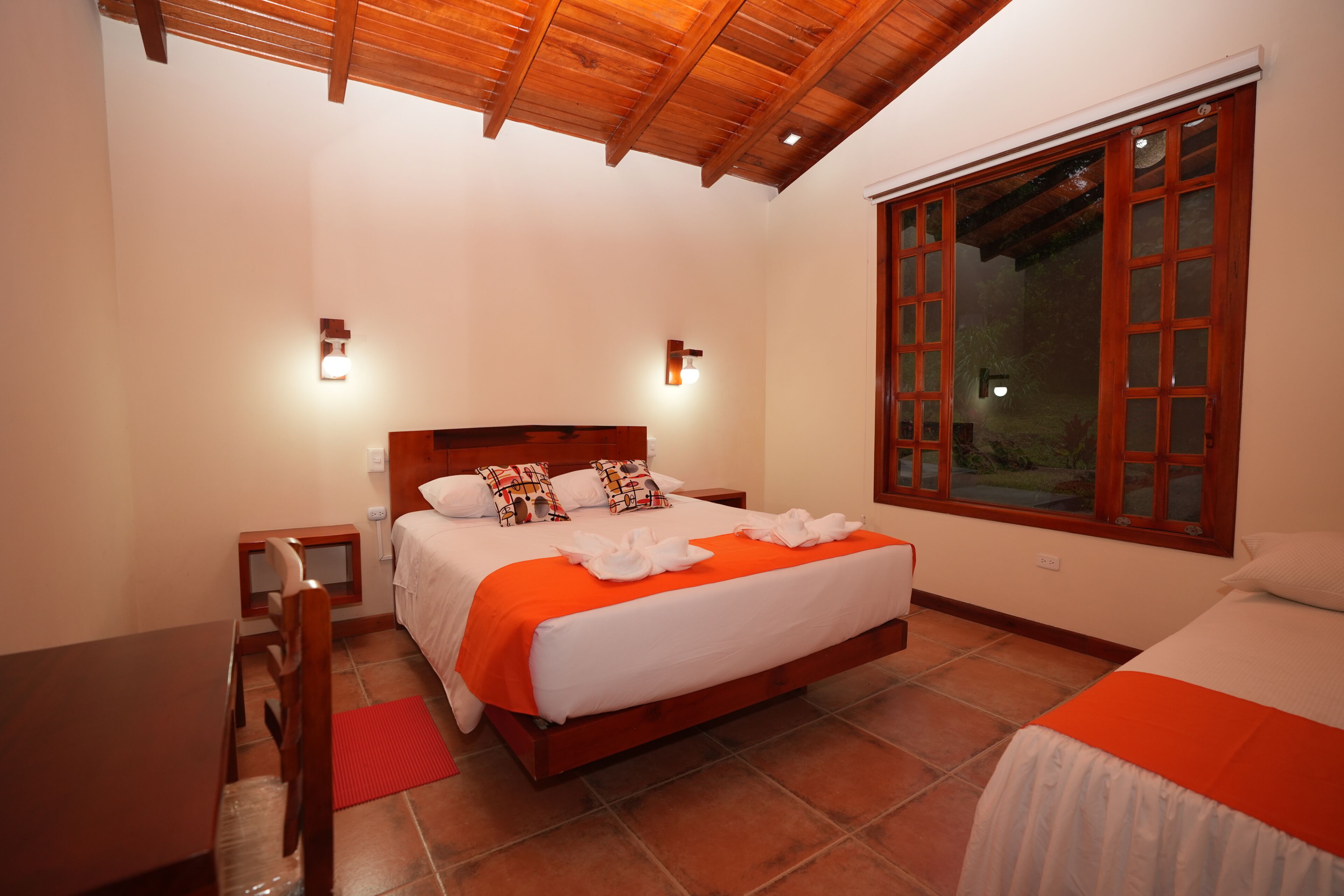 Un dormitorio con techo de madera, una cama grande con colcha naranja, dos mesitas de noche y una ventana con contraventanas de madera.