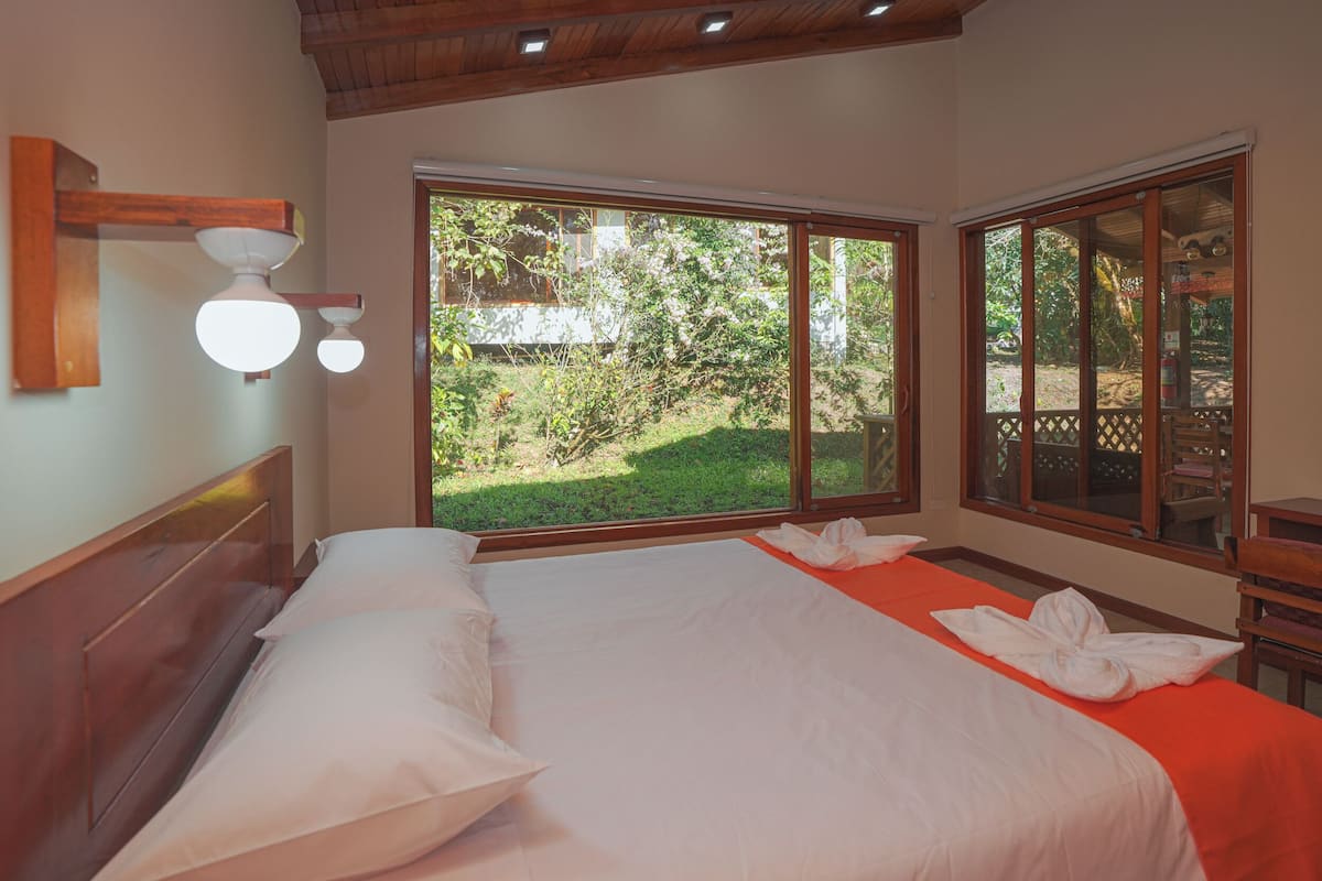 A bedroom with a large bed, a wooden headboard, a window with a view of greenery, and a ceiling light fixture.