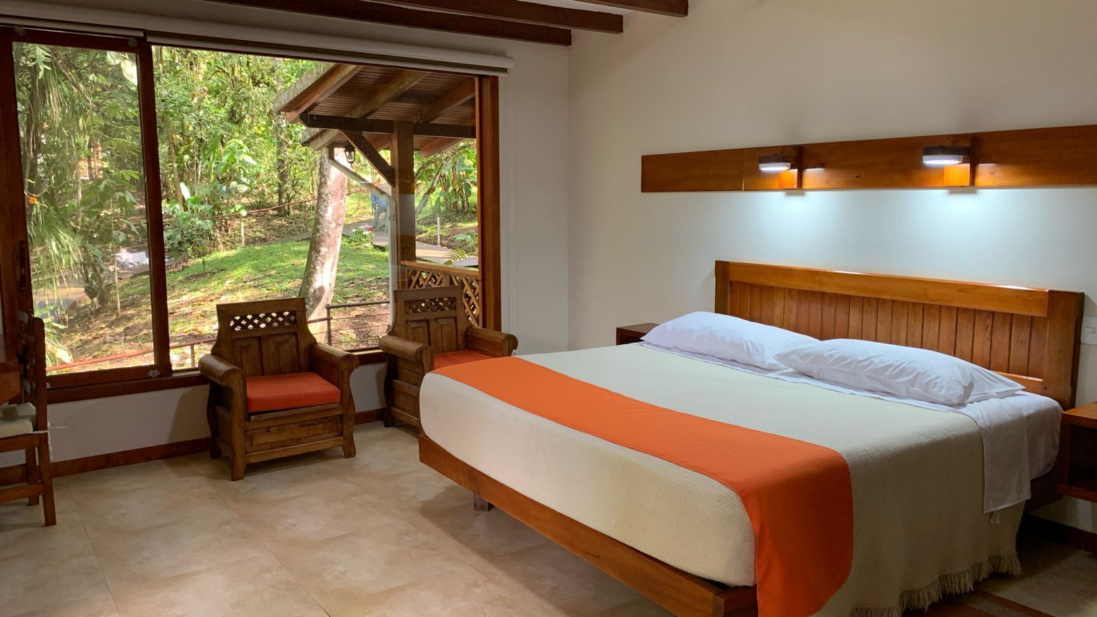A bedroom with a bed, wooden headboard, two nightstands, a wooden chair, and a large window with a view of greenery.