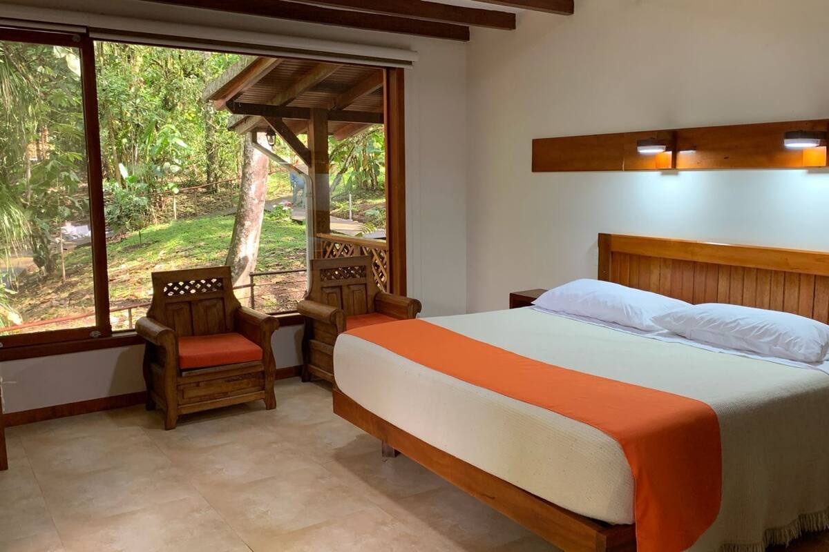 A bedroom with a bed, wooden headboard, two nightstands, a wooden chair, and a large window with a view of greenery.