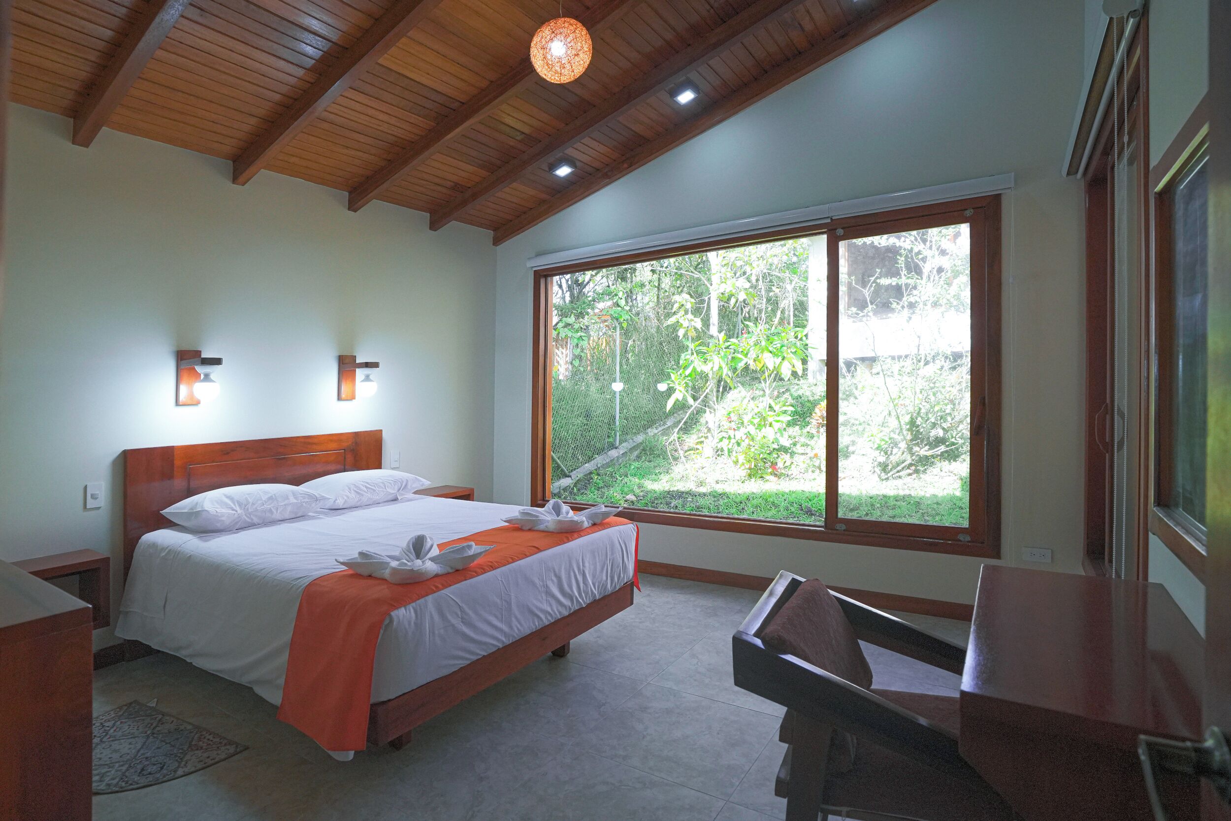 Une chambre avec un plafond en bois, une grande fenêtre, un lit recouvert de draps blancs et un bureau.