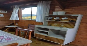 A wooden cabin interior with a dining table, chairs, a small round table, a white cabinet with shelves, and a window with curtains.