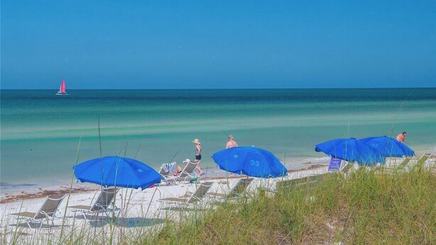 Een strand met ligbedden en parasols, een zeilboot in de verte, en een heldere blauwe lucht.