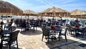 Een terras buiten met tafels en stoelen, rieten parasols en een zwembad op de achtergrond.