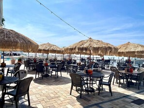 Eine Terrasses mit Tischen, Stühlen und einem Pool im Hintergrund, beschattet durch Strohdecken.