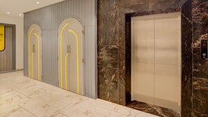 Un ascenseur avec un mur en marbre, une porte grise agrémentée de touches de jaune et une caméra de surveillance bien visible.