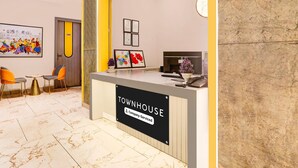 Un comptoir d’accueil portant l’inscription « TOWNHOUSE » et un panneau indiquant « Service aux entreprises ». On peut y voir des certificats encadrés accrochés au mur, une peinture colorée et une petite table avec des chaises.