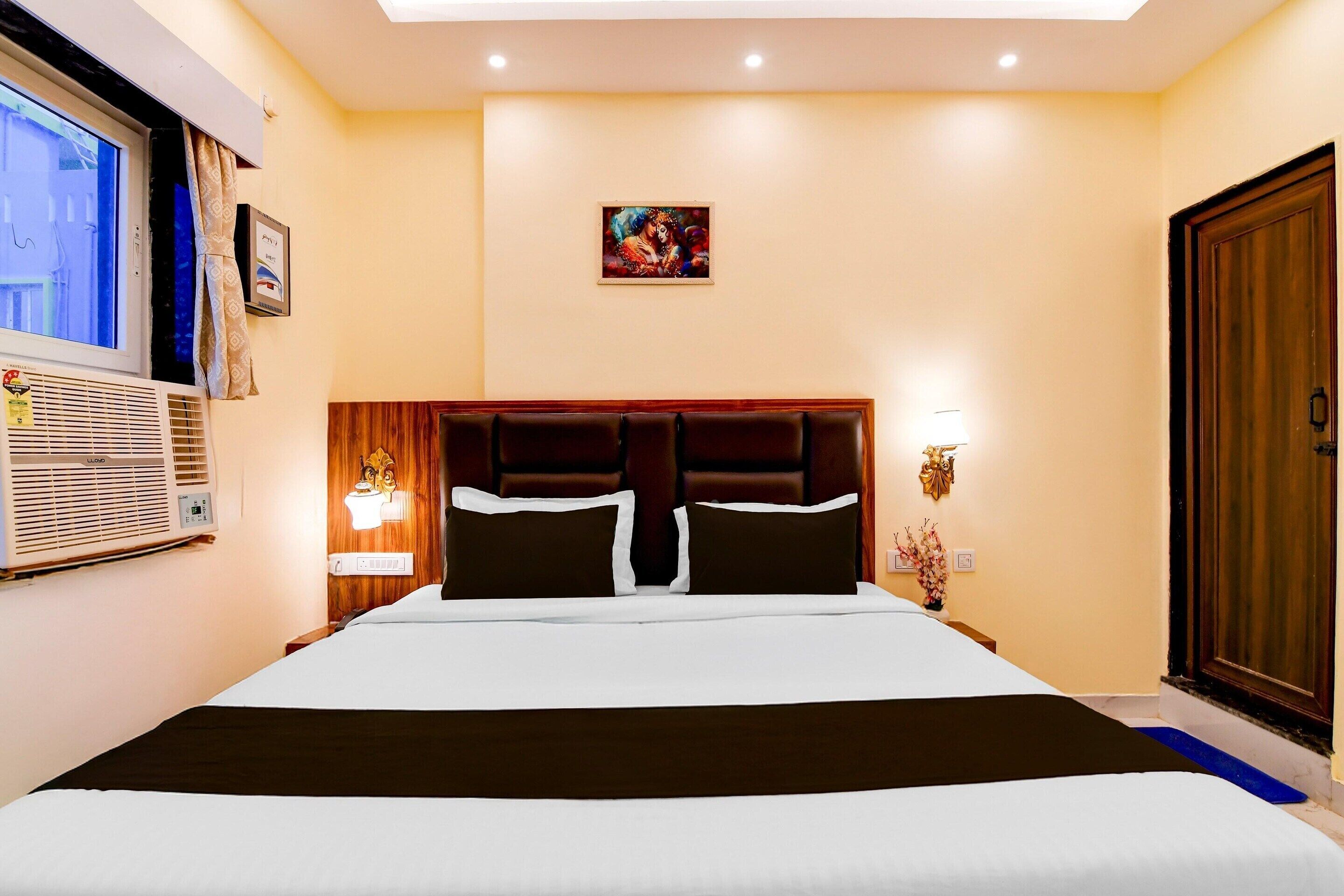 A hotel room with a large bed, a headboard, a window with blinds, a wall-mounted air conditioner, a framed picture, and a door.