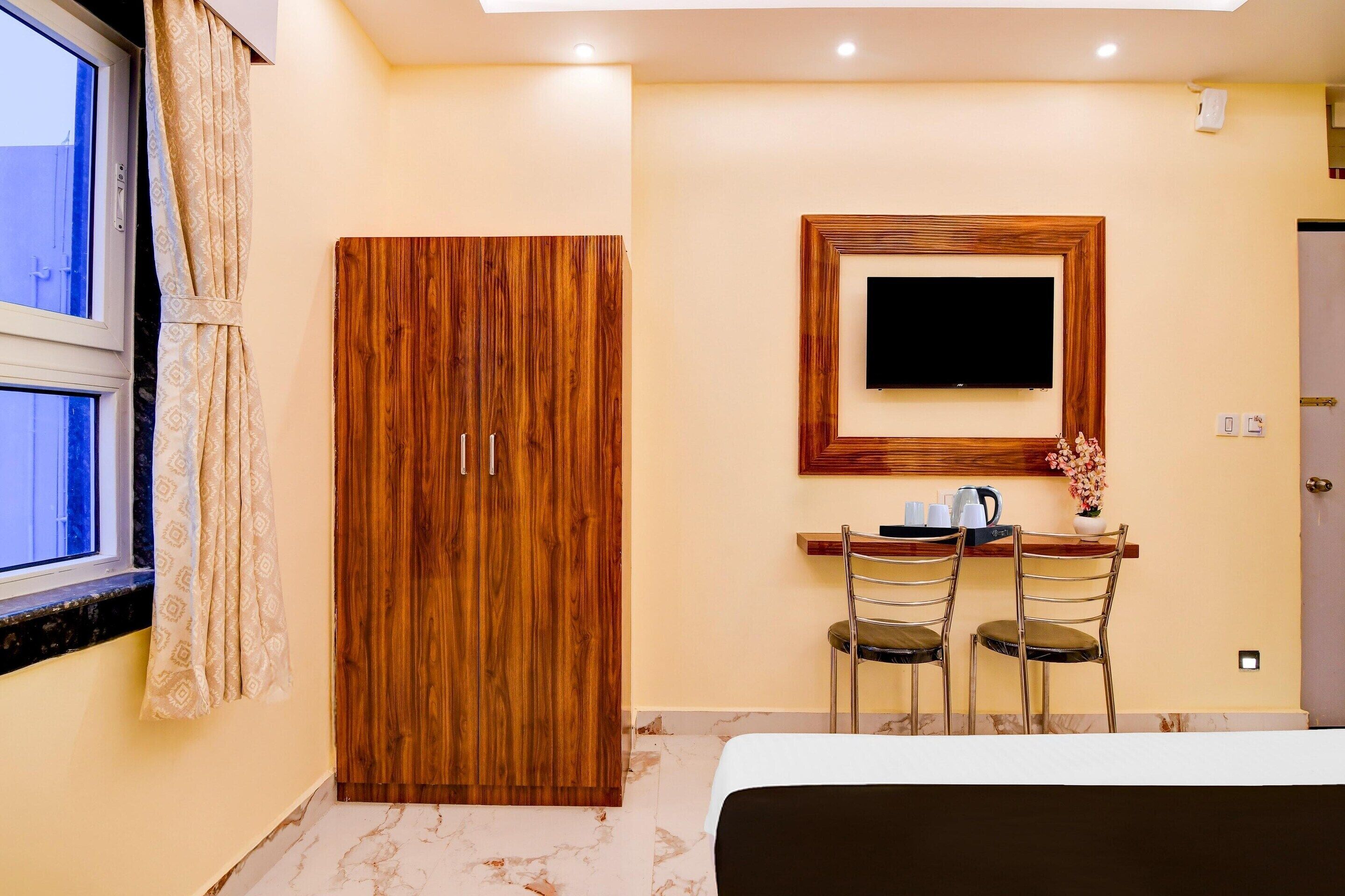 A hotel room with a wooden wardrobe, a TV mounted on the wall, a desk with chairs, and a bed.