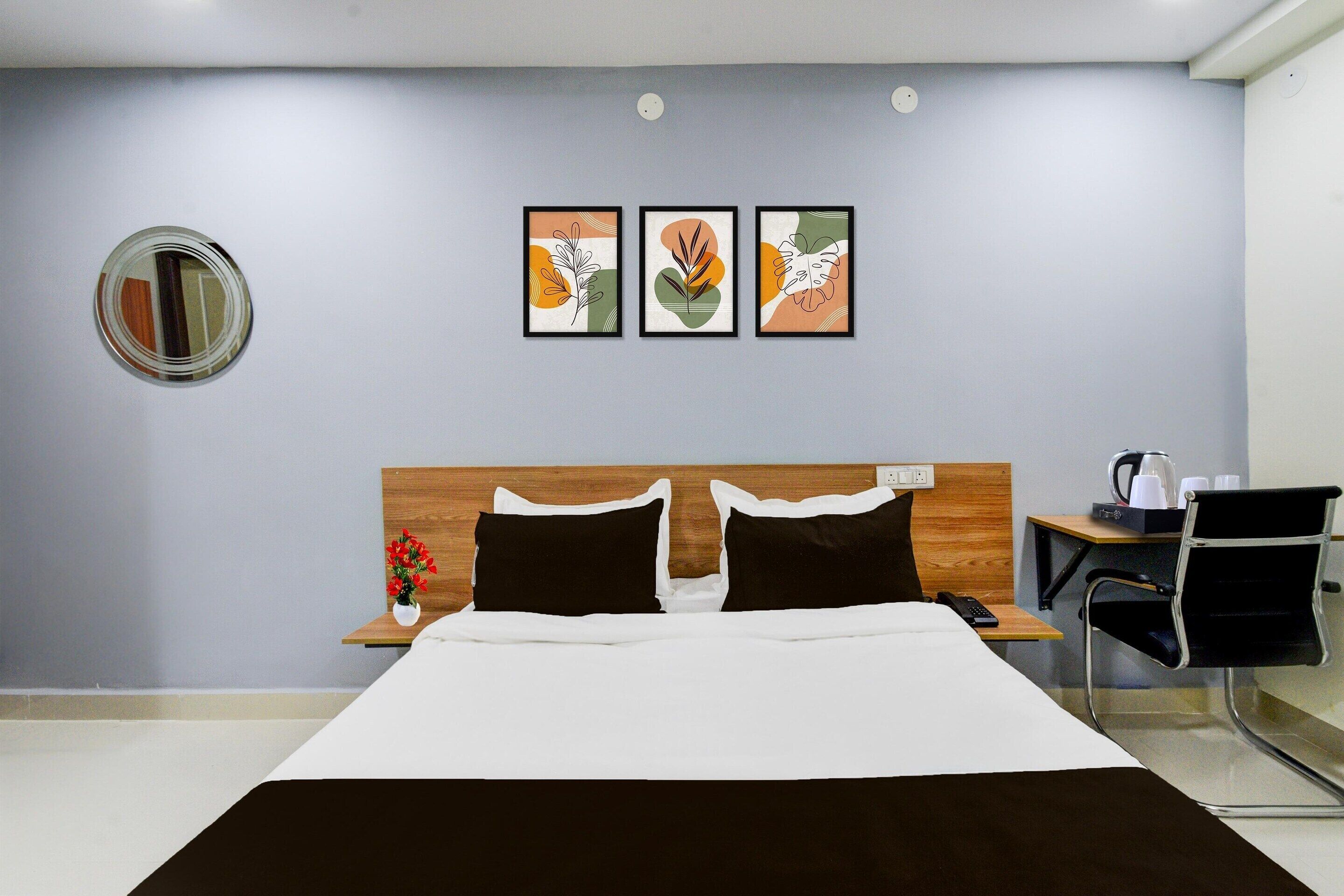 A hotel room with a bed, a desk with a chair, a coffee maker, and three framed artworks on the wall.