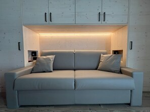 A modern, light-colored sofa with a built-in light feature above it, against a wooden wall. - Bormio Apartments al pradais giardino ovest (Bormio)
