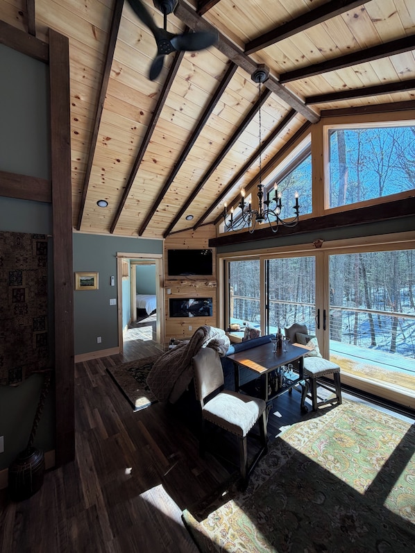A spacious room with a high ceiling, wooden beams, a dining area with a table and chairs, and large windows offering a view of the outdoors.