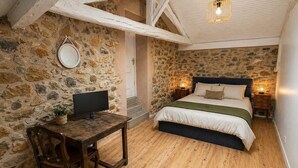 A bedroom with a stone wall, wooden floor, a bed with white bedding, a bedside table with a lamp, and a TV on a wooden table. - La Petite Maison - Spa - Games room - Air conditioning - Garden (Arguenos)