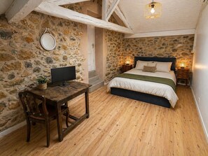 A bedroom with a stone wall, wooden floor, a bed with white bedding, a bedside table with a lamp, and a TV on a wooden table. - La Petite Maison - Spa - Games room - Air conditioning - Garden (Arguenos)
