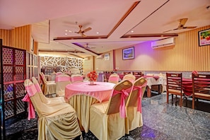 Un salón de banquetes con mesas redondas, sillas con cintas rosas, ventiladores de techo y un equipo de aire acondicionado.