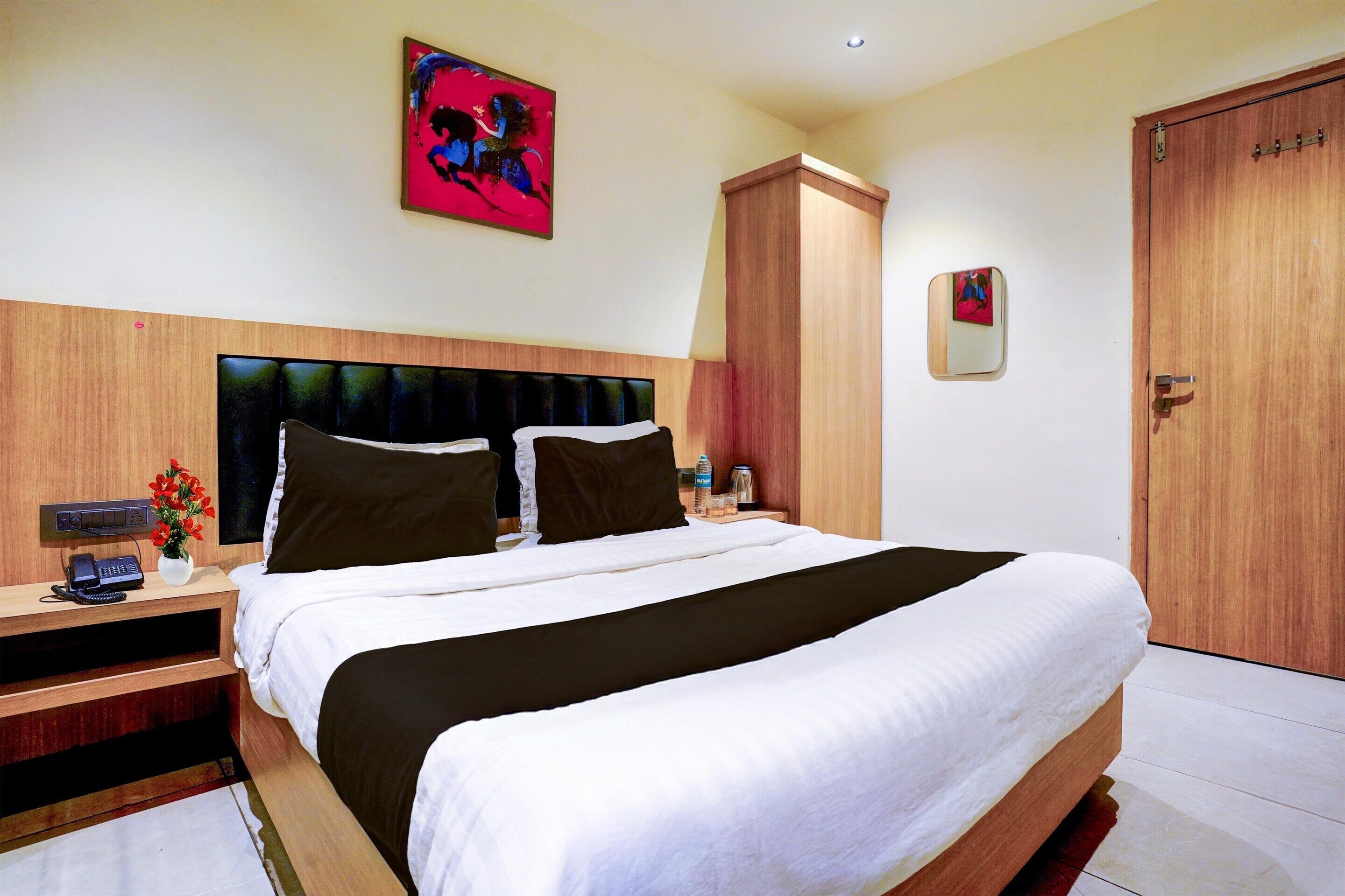 A hotel room with a large bed, a wooden headboard, a nightstand with a phone and a vase of flowers, and a painting on the wall.
