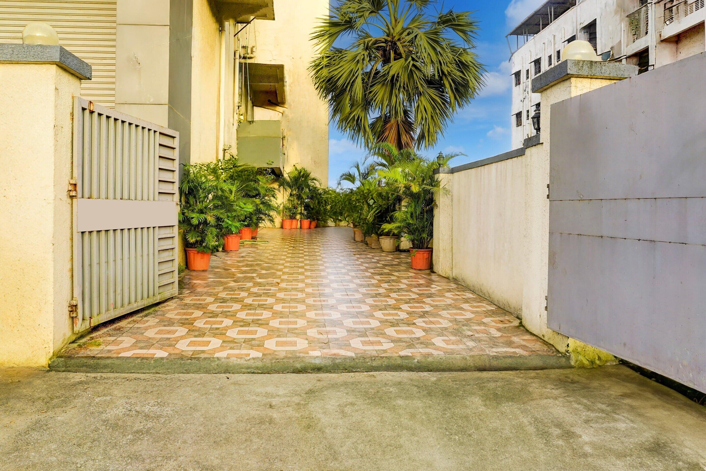 A narrow pathway with patterned tiles leading to a gated entrance, surrounded by potted plants and a palm tree.