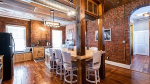 A rustic kitchen with wooden cabinets, a large island with chairs, and a brick wall. - New! The Old Timer - 5BR/4.5BA - Hot Tub! Historic! (Louisville)