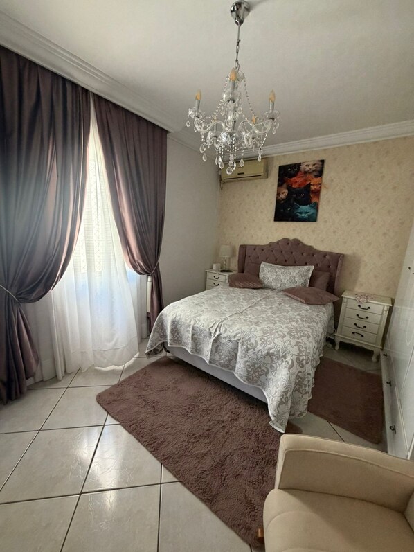 A bedroom with a bed, a nightstand, a dresser, a chair, a chandelier, and a window with curtains.