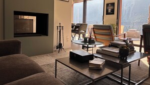 Un salón con sofá, mesa de centro, sillas, chimenea y ventanas amplias con vistas.