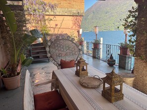Ein Balkon mit Korbmöbeln, Topfpflanzen und dekorativen Laternen mit Blick auf ein Gewässer und Hügel.