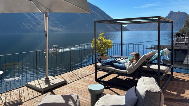 Un balcón con vistas a un lago y montañas, una silla de descanso, una mesita y una planta en maceta.