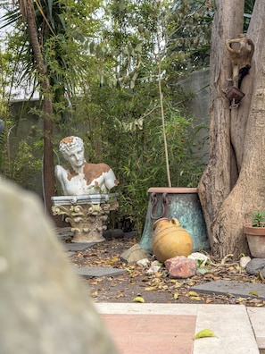 Ein Garten mit einer Statue, einem Baum und verschiedenen Kübelpflanzen.