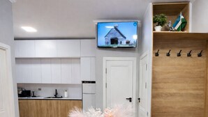 Una sala de estar moderna con una mesa de centro redonda de madera, un jarrón blanco con flores, un televisor de pantalla plana montado en la pared y un perchero de madera con ganchos.