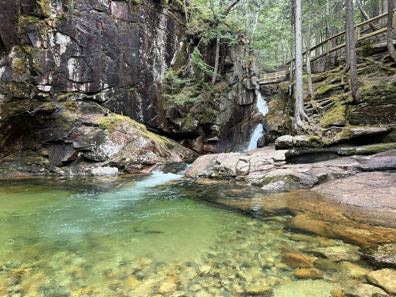 Eine natürliche Felslandschaft mit einem klaren Bach, einer Holzbrücke und einem Wasserfall.