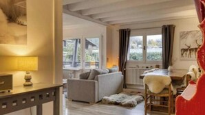 Una sala de estar acogedora con sofá, una mesa y una silla. Hay ventanas con cortinas, un radiador y vistas al exterior.