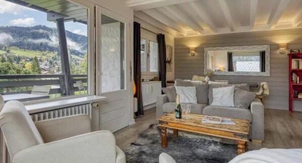A cozy living room with a sofa, a coffee table, and a view of a mountainous landscape through large windows. - Charming apartment in the heart of Megève with terrace and mountain view (Combloux)