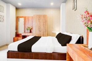 A hotel room with a bed, a wooden nightstand, a mirror, a vase with flowers, and a wall with a textured pattern. - Classic With Balcony Beltola Tiniali Pratishtha (Guwahati)
