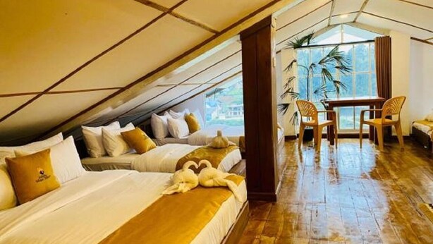 Ein Hotelzimmer mit schrägverdeckter Decke, zwei Betten mit weißen Leinen, Holzboden, einer Essecke mit Tisch und Stühlen sowie Blick durch ein großes Fenster.